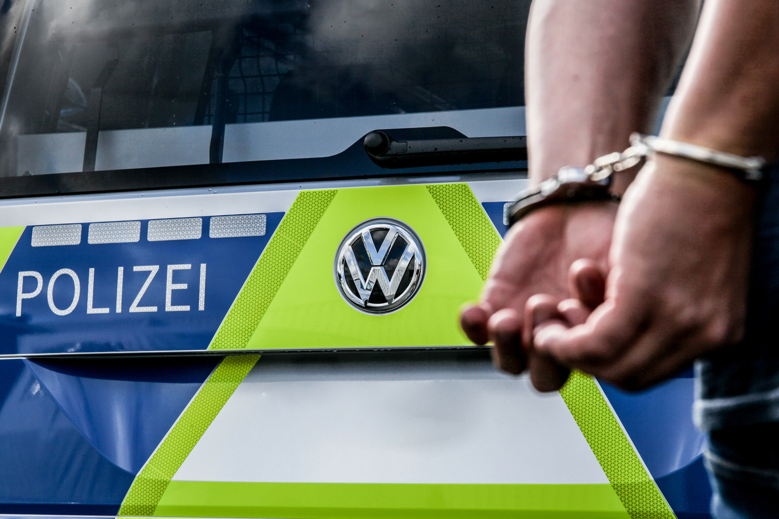 Vor einem Polizeiauto steht eine Person in Handschellen.