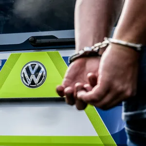 Vor einem Polizeiauto steht eine Person in Handschellen.