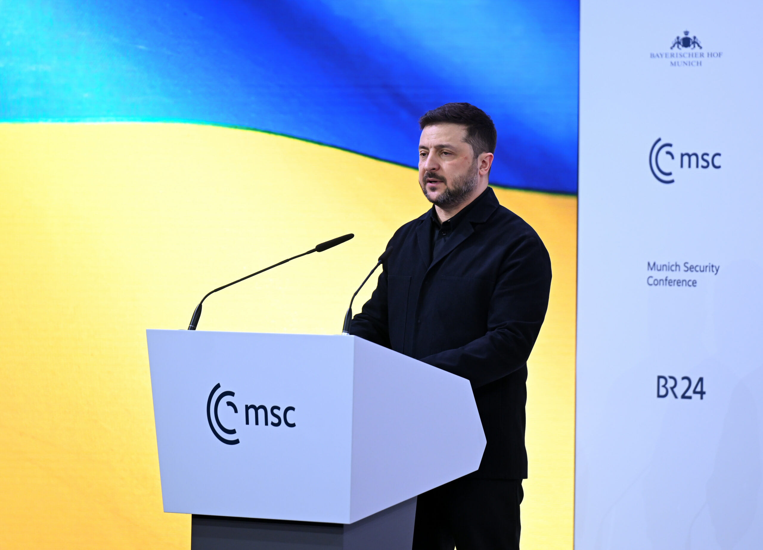 Mann mit Bart an Rednerpult vor gelb-blauer Wand