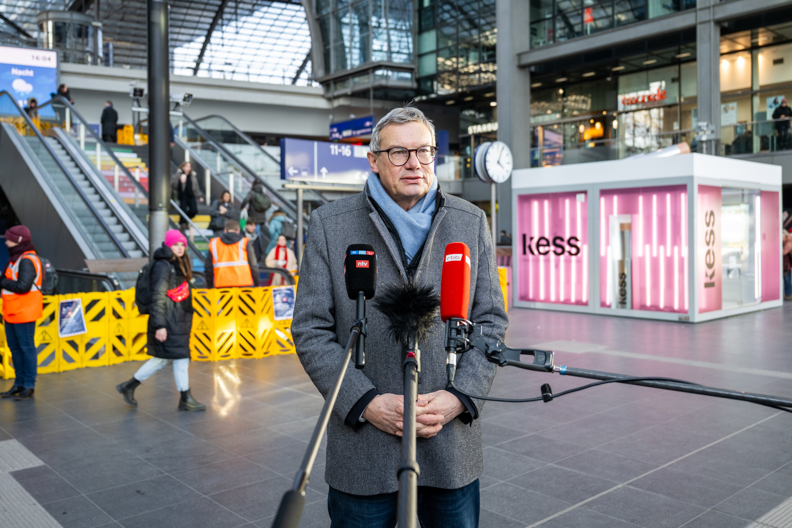 Mann steht vor Mikrofonen, im Hintergrund Rolltreppen