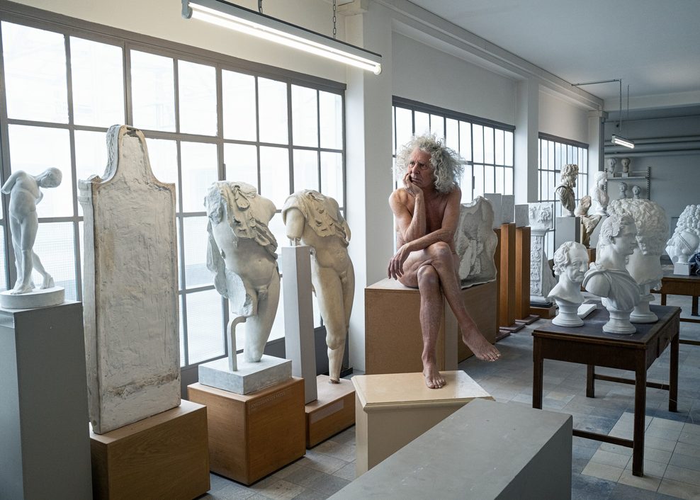 Rainer Langhans sitzt neben mehreren Statuen auf einem Sockel.