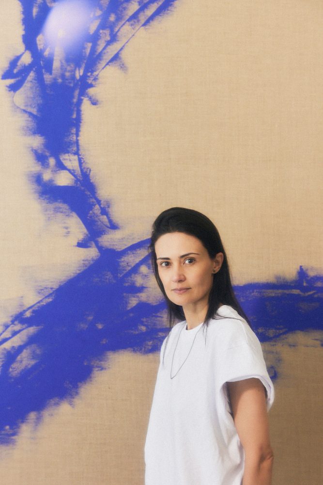 Sarice Brudet vor einer hellbraunen/blauen Wand. Sie hat dunkle Haare und trägt ein weißes T-Shirt.