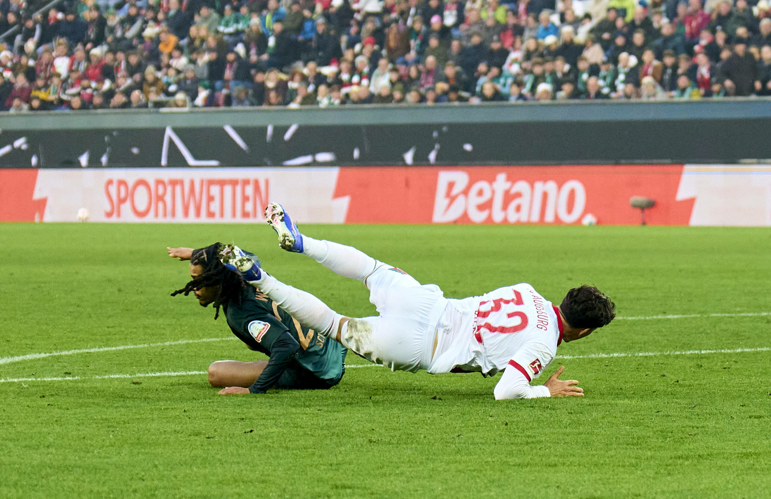 Isaac Schmidt von Werder Bremen grätscht einen Gegenspieler um. Im Hintergrund läuft Bandenwerbung für den Wettanbieter Betano.