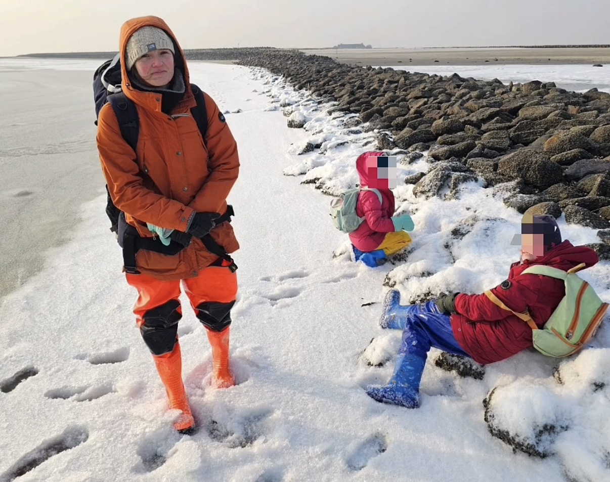 Eine Frau und ihre zwei Kinder stehen und sitzen am Wattenmeer. Die Gesichter der Kinder sind gepixelt.