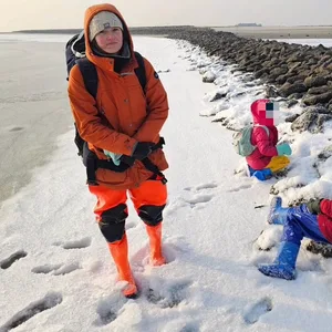 Eine Frau und ihre zwei Kinder stehen und sitzen am Wattenmeer. Die Gesichter der Kinder sind gepixelt.