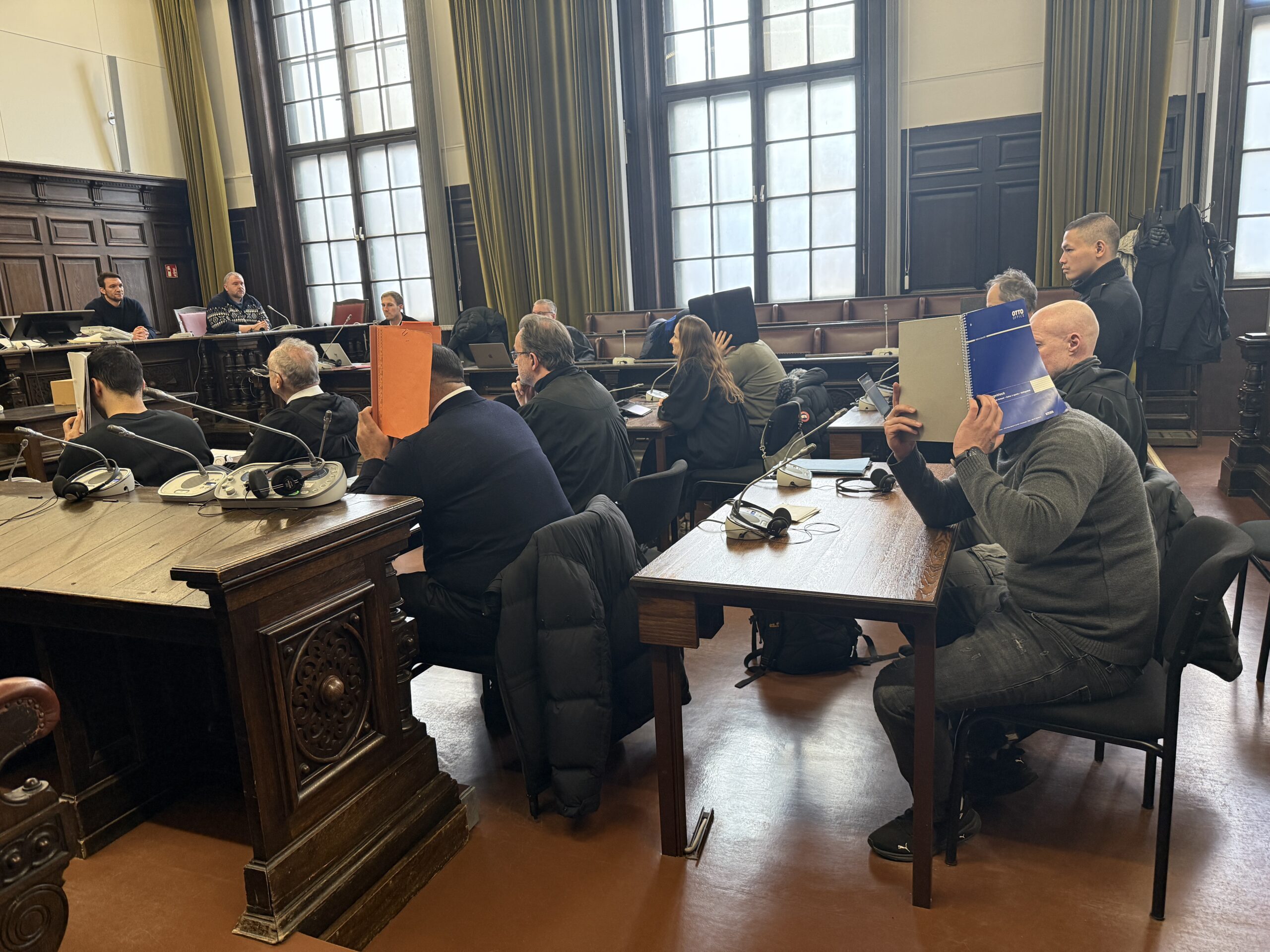 Die Angeklagten sitzen neben ihren Verteidiger:innen und halten sich Mappen vor das Gesicht.