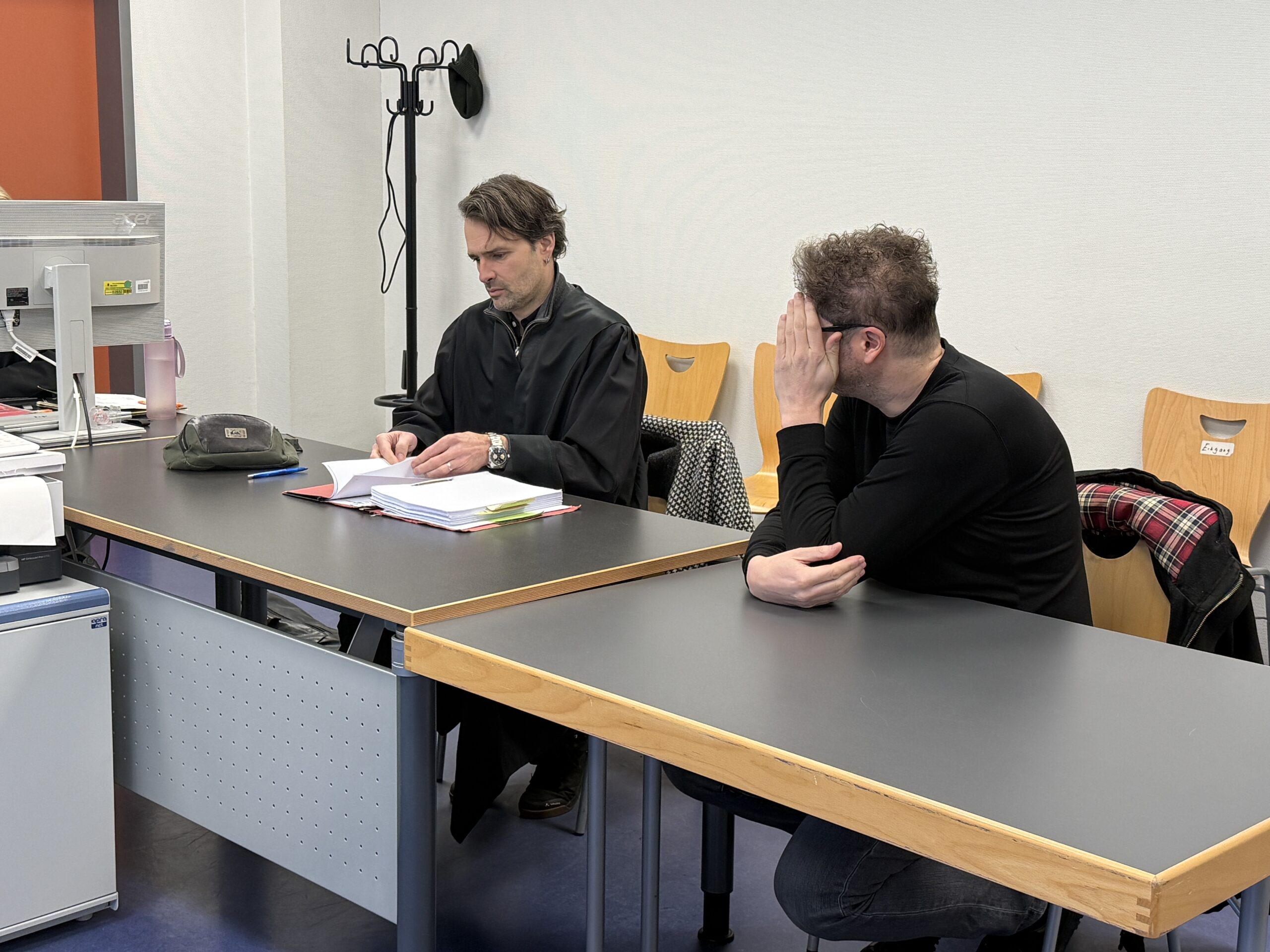 Der Angeklagte sitzt neben seinem Verteidiger. Er hält sich die Hand vor das Gesicht und dreht den Kopf weg.