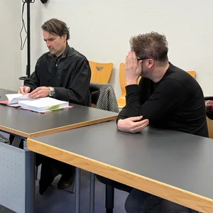 Der Angeklagte sitzt neben seinem Verteidiger. Er hält sich die Hand vor das Gesicht und dreht den Kopf weg.