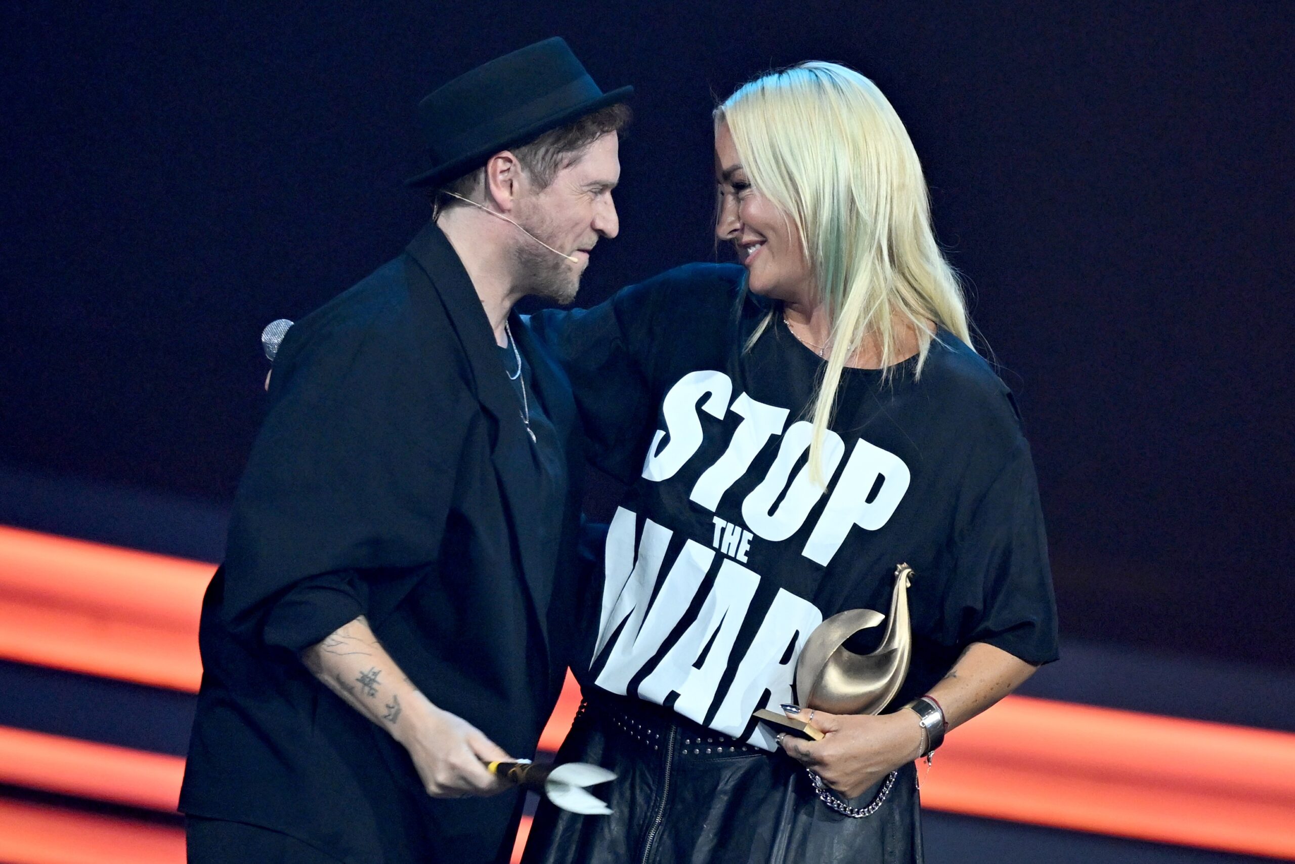 Sie trägt ein T-Shirt, auf dem „Stop The War“ steht, hält den Preis in der Hand und umarmt Johannes Oerding