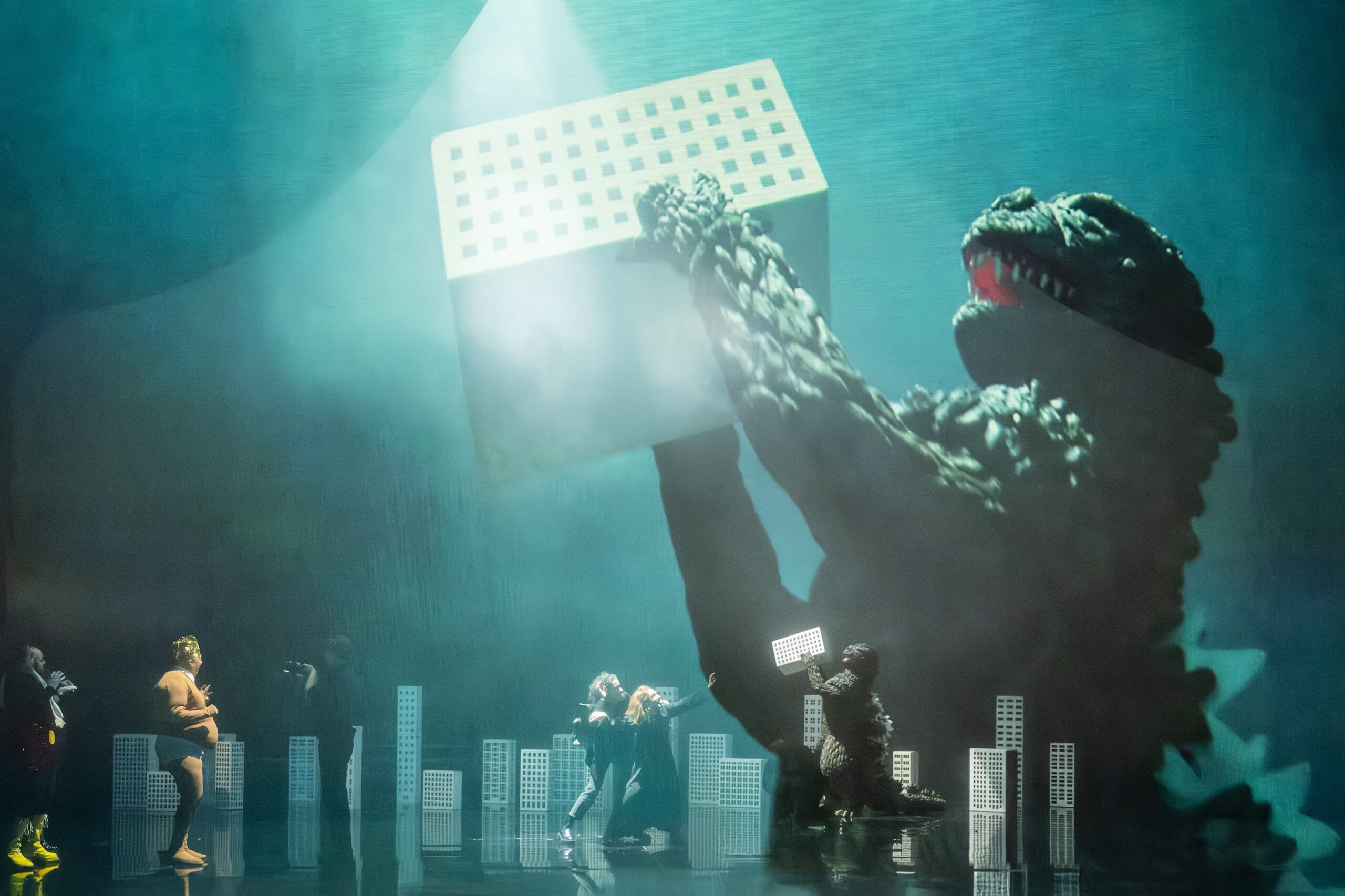 Szene: Man sieht ein paar Menschen auf der Bühne und auch das Monster, das ein Hochhaus-Modell in den Händen hält. Es ist riesig auf eine Leindwand projiziert