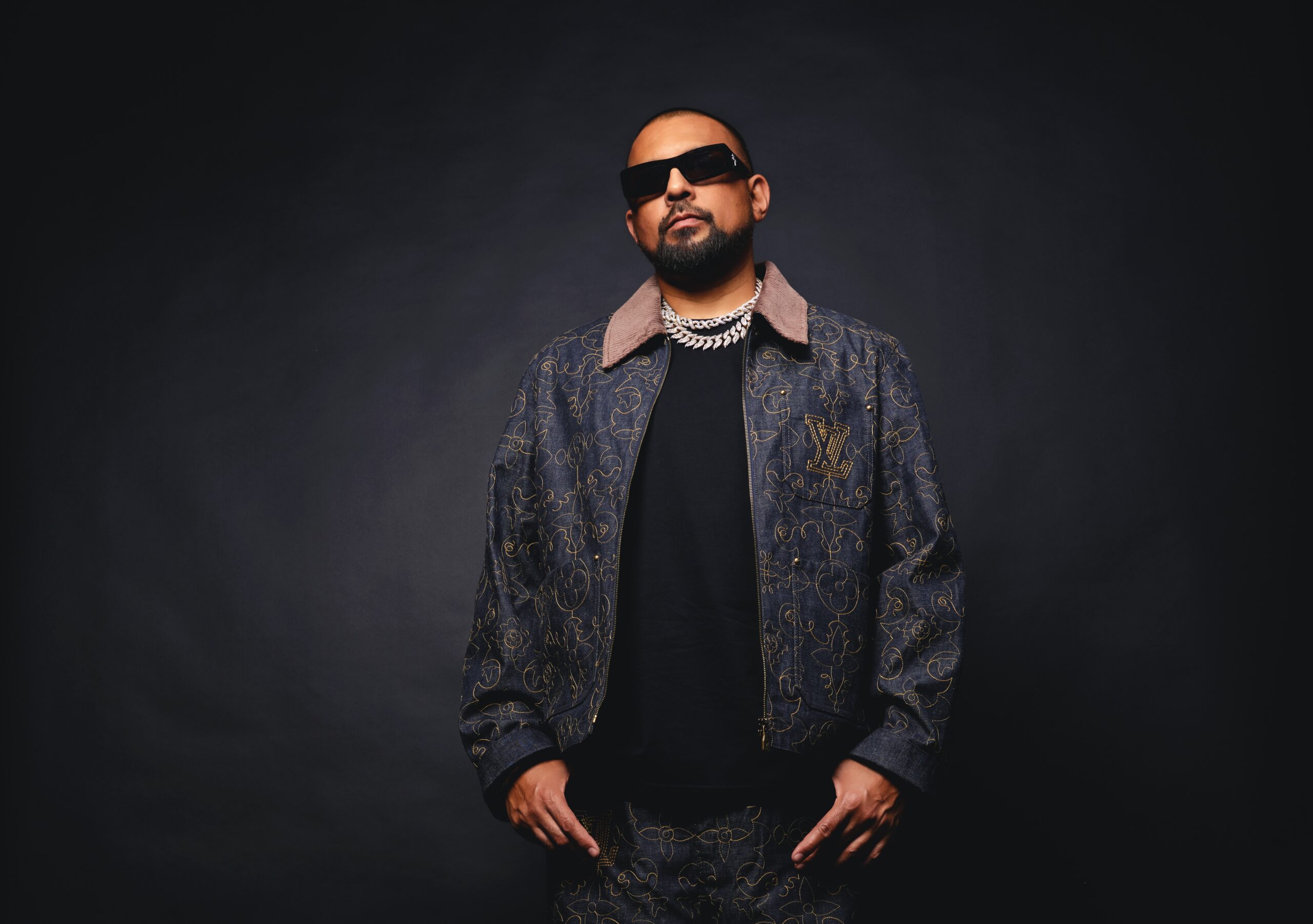 Er steht vor einer dunklen Wand, trägt Sonnenbrille, hat die Daumen in den Hosentaschen und blickt ernst in die Kamera