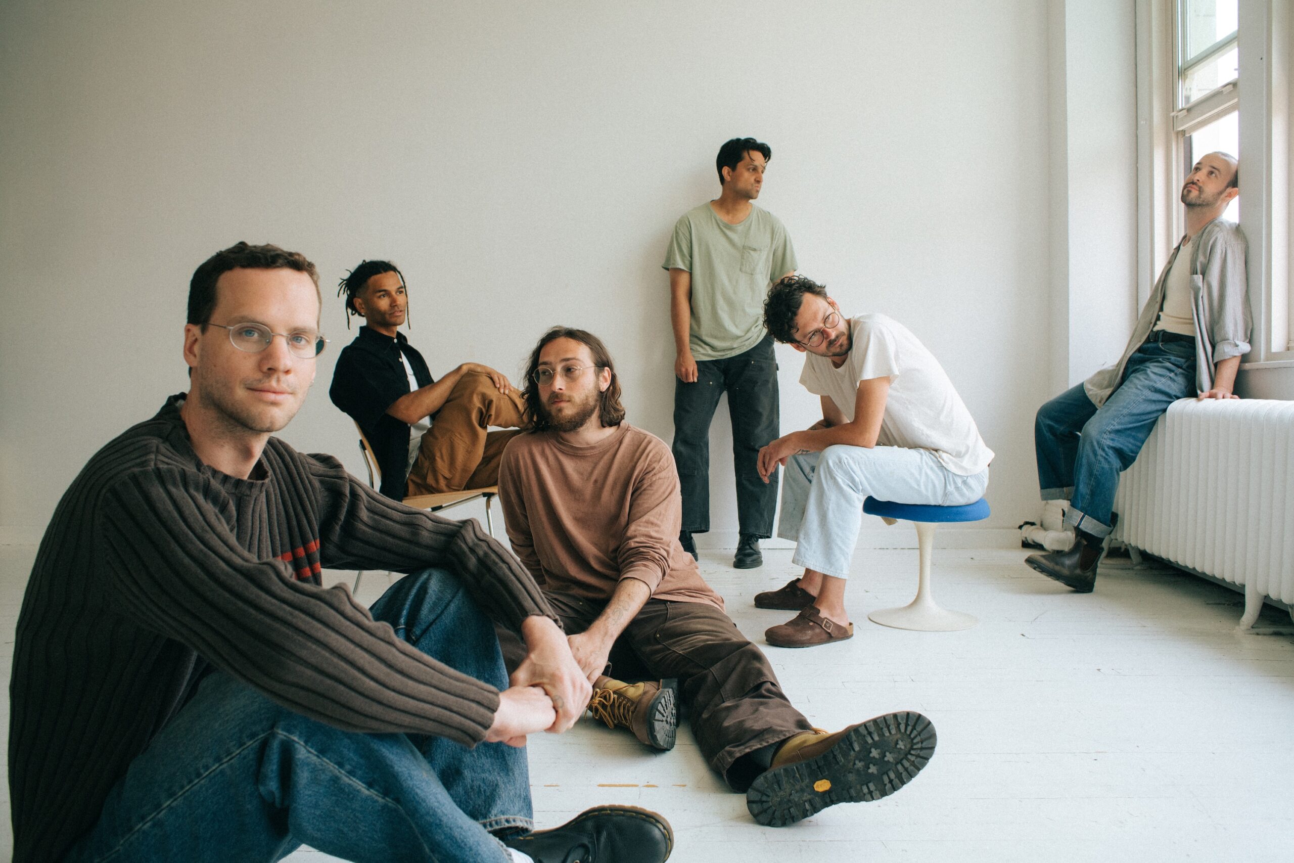 Die Band – sechs Männer - in einem weißen Raum, sie sitzen auf dem Boden, auf Stühlen, auf einem Heizkörper oder lehnen an der Wand