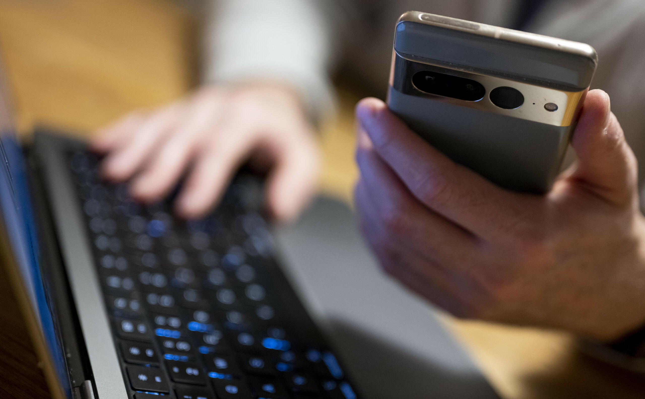 Eine Person sitzt am Laptop und hält ein Handy in der Hand.