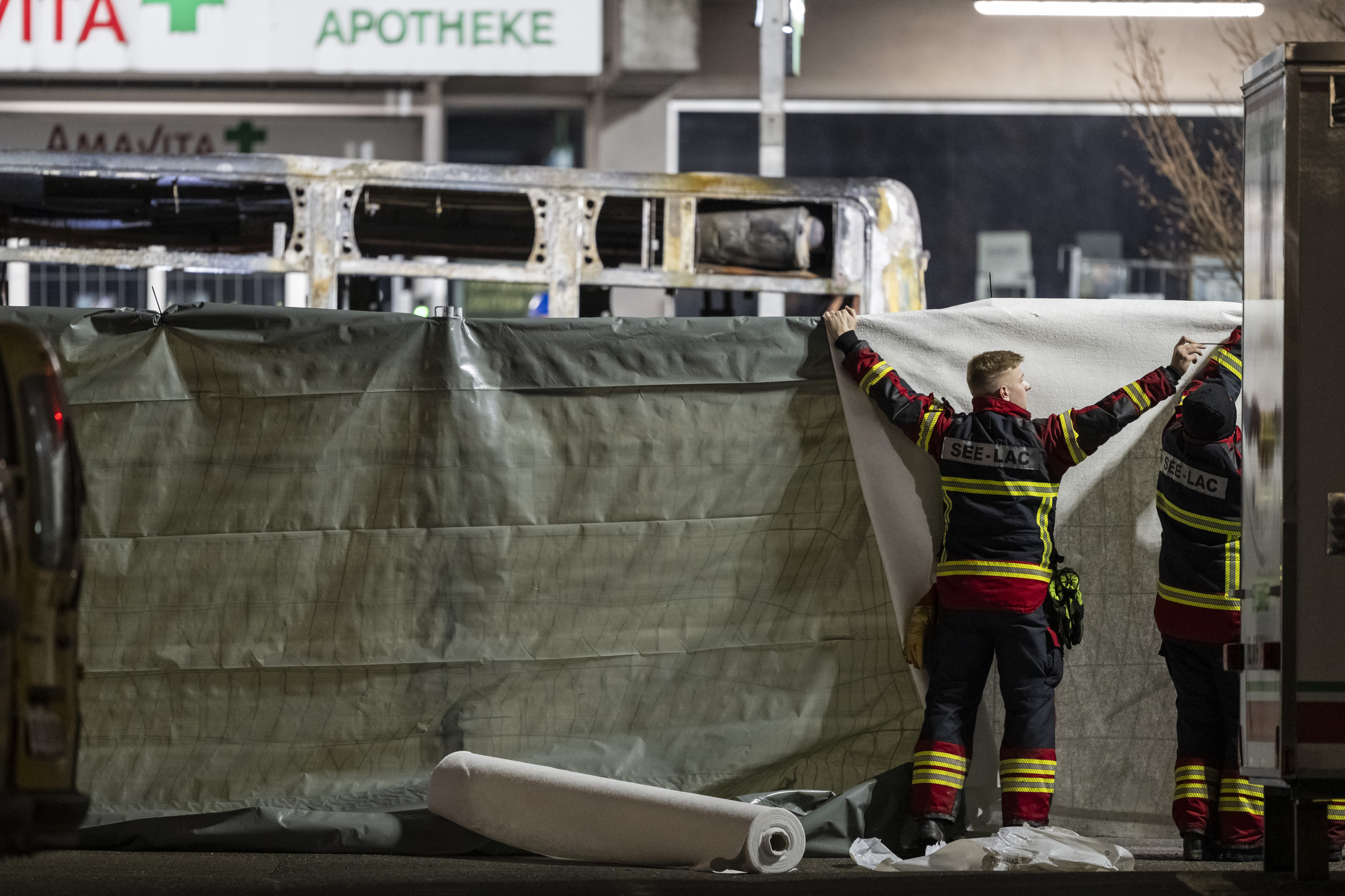 Feuerwehrleute verdecken den Bereich vor der verkohlten Hülle eines ausgebrannten Linienbusses.