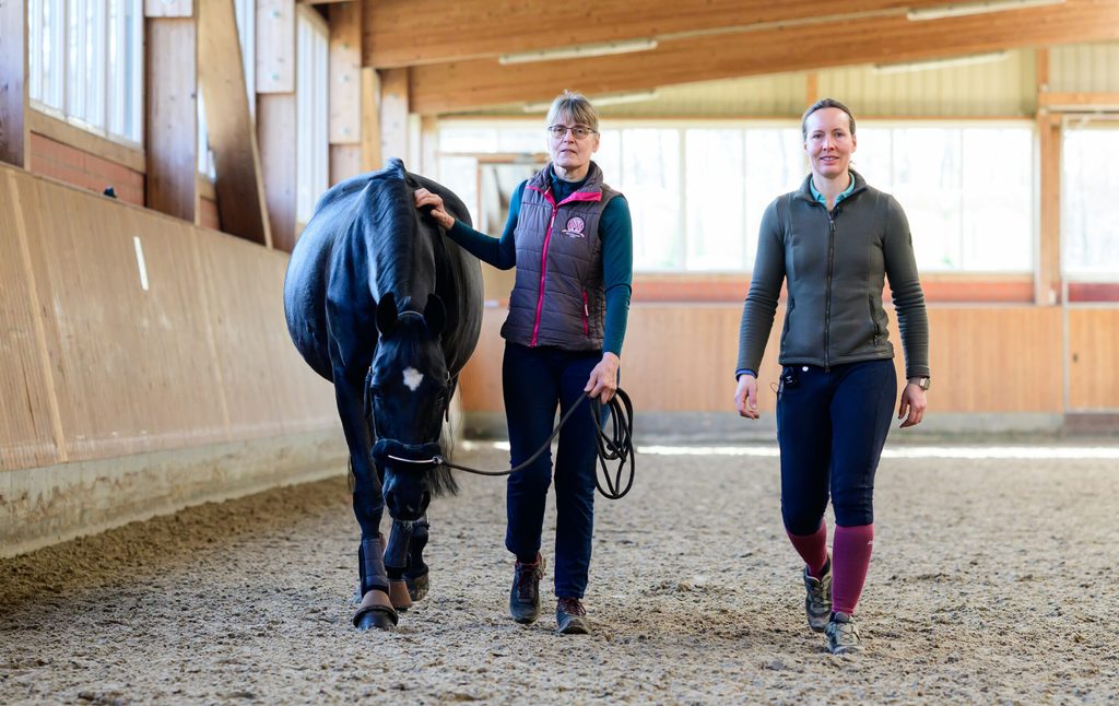 Andrea Engeler-Bading (r), Dressurreiterin und Besitzerin des Hofes Fehmbusch, und Imke Steffan, Mitarbeiterin, gehen zusammen mit der Hannoveraner Stute Carmina durch eine Reithalle.