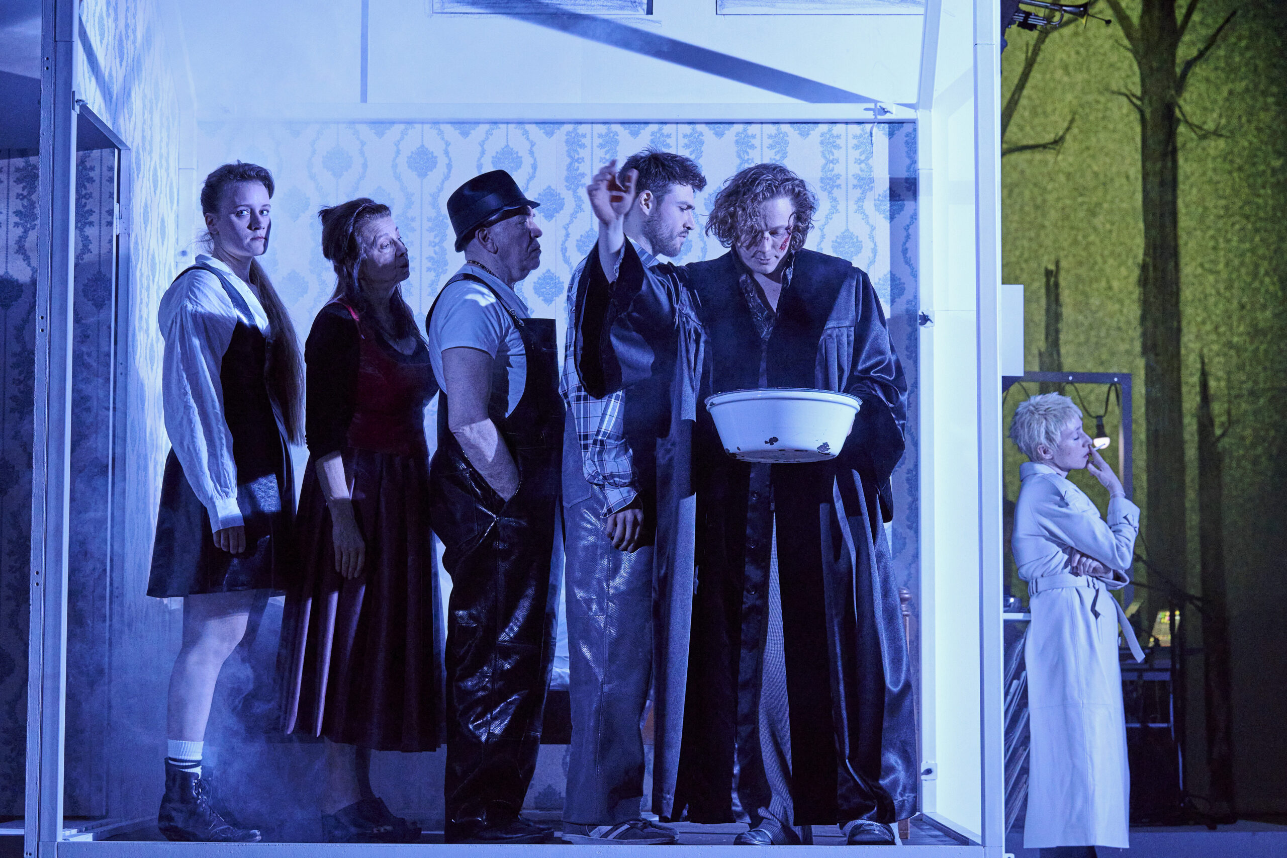 Szene aus dem Stück, auf der Bühne stehen fünf Personen in einem Raum, eine weitere Person steht rechts davor
