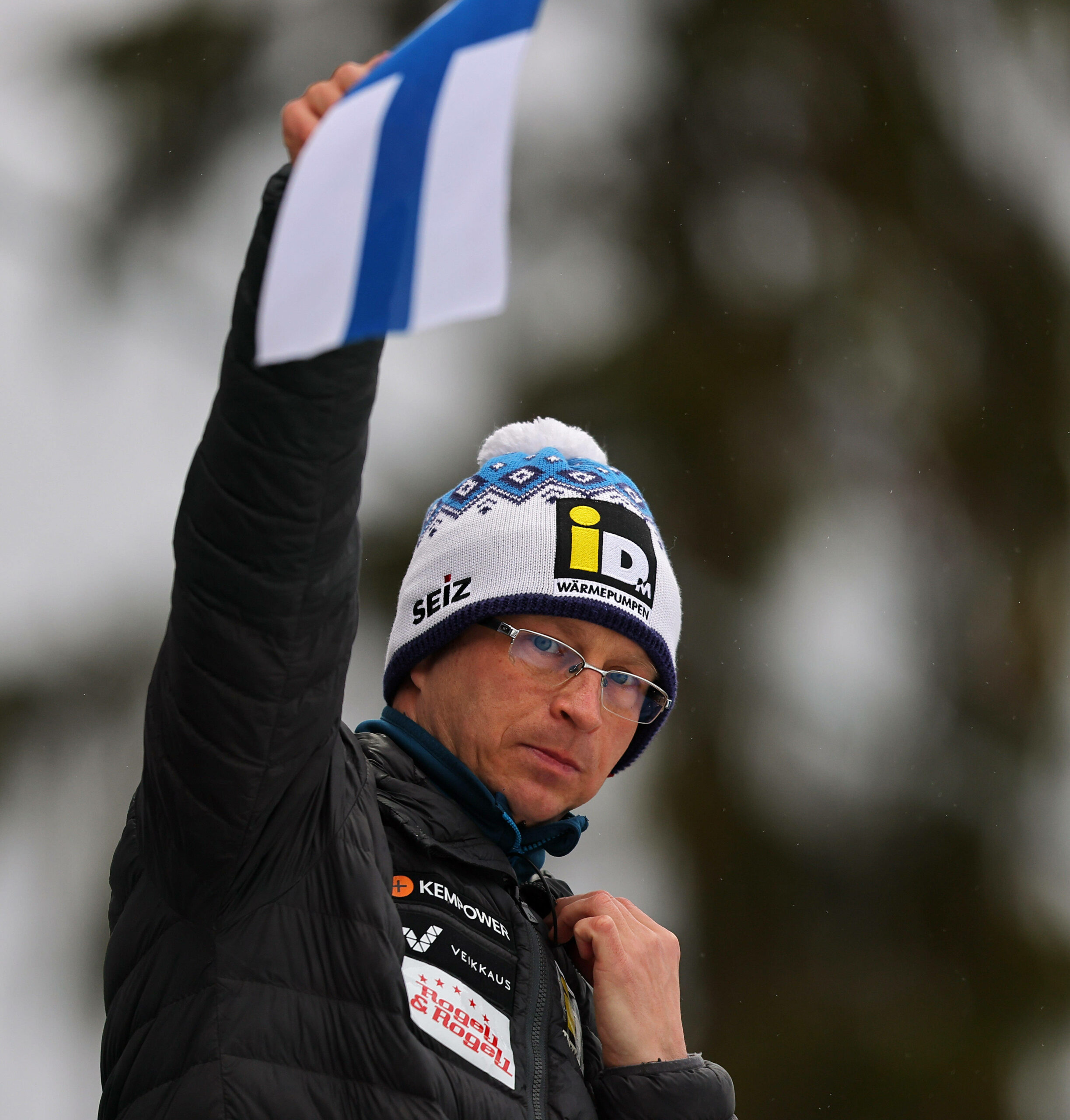 Igor Medved hält die finnische Fahne als Startsignal für einen Skisprung-Athleten hoch.