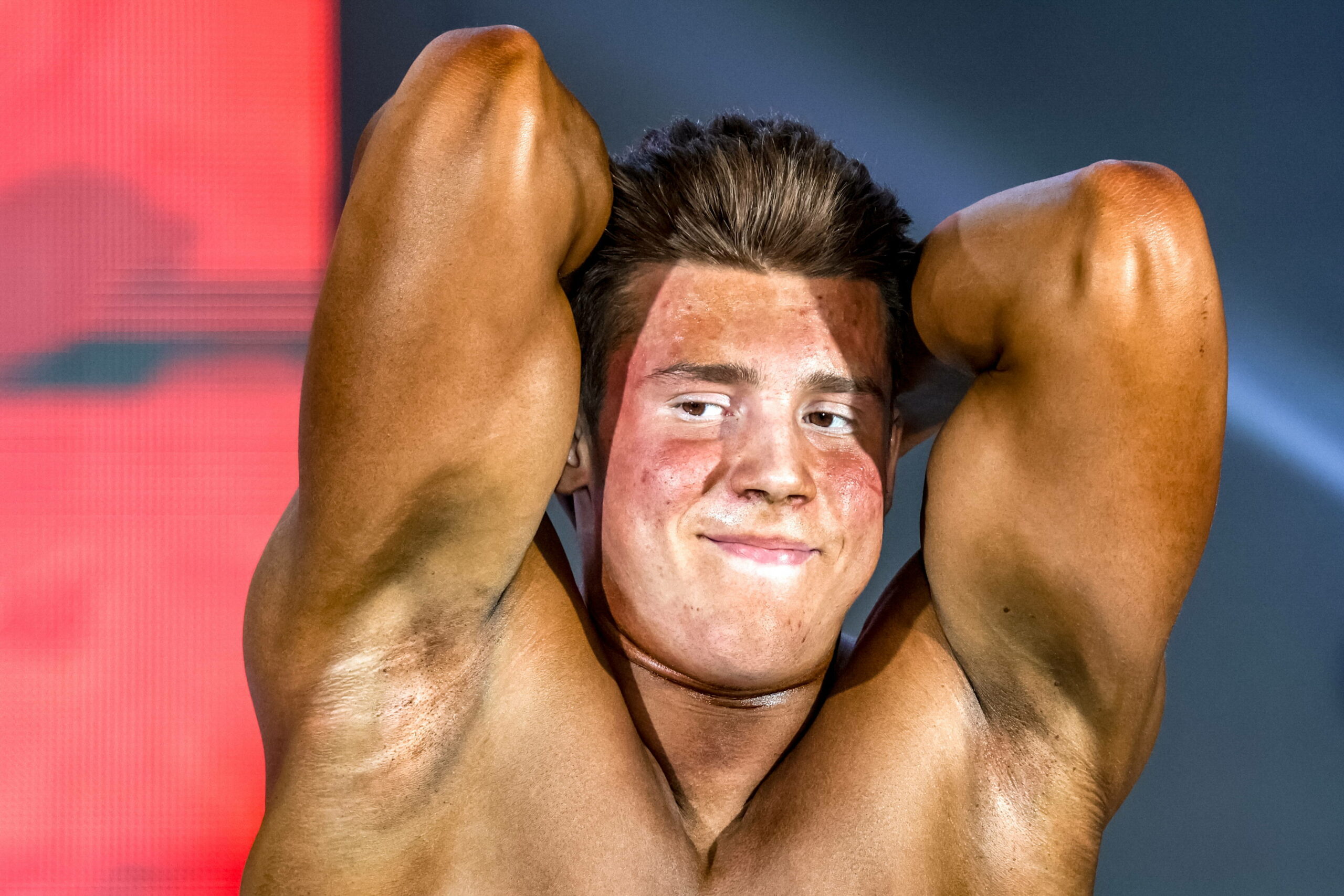 Ein Mann hat die Hände hinter dem roten Kopf. Er spannt seine Muskeln an. Im Gesicht wirkt er unentspannt.