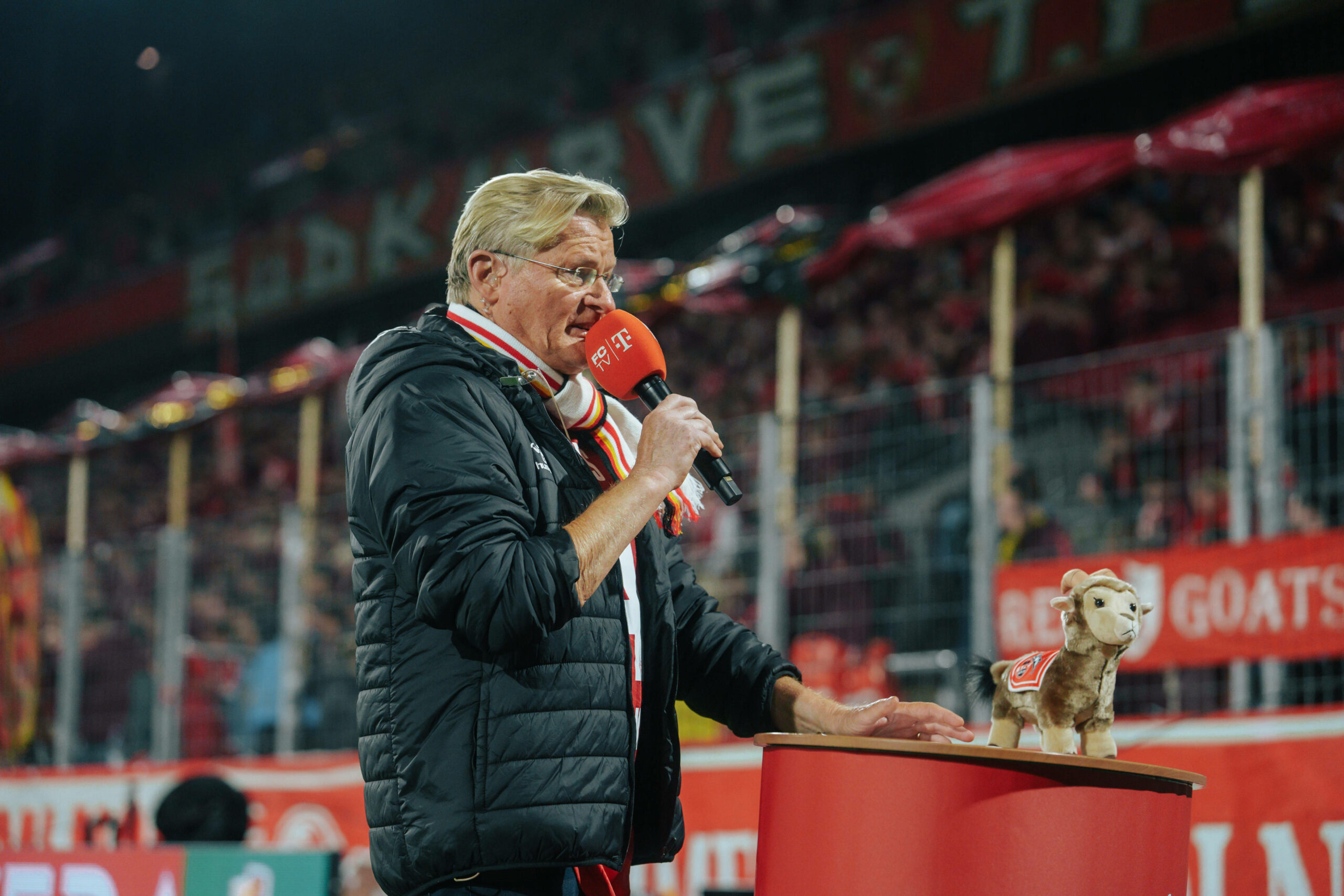 Michael Trippel spricht in ein Mikrofon. Im Hintergrund ist die Fankurve des 1. FC Köln zu sehen