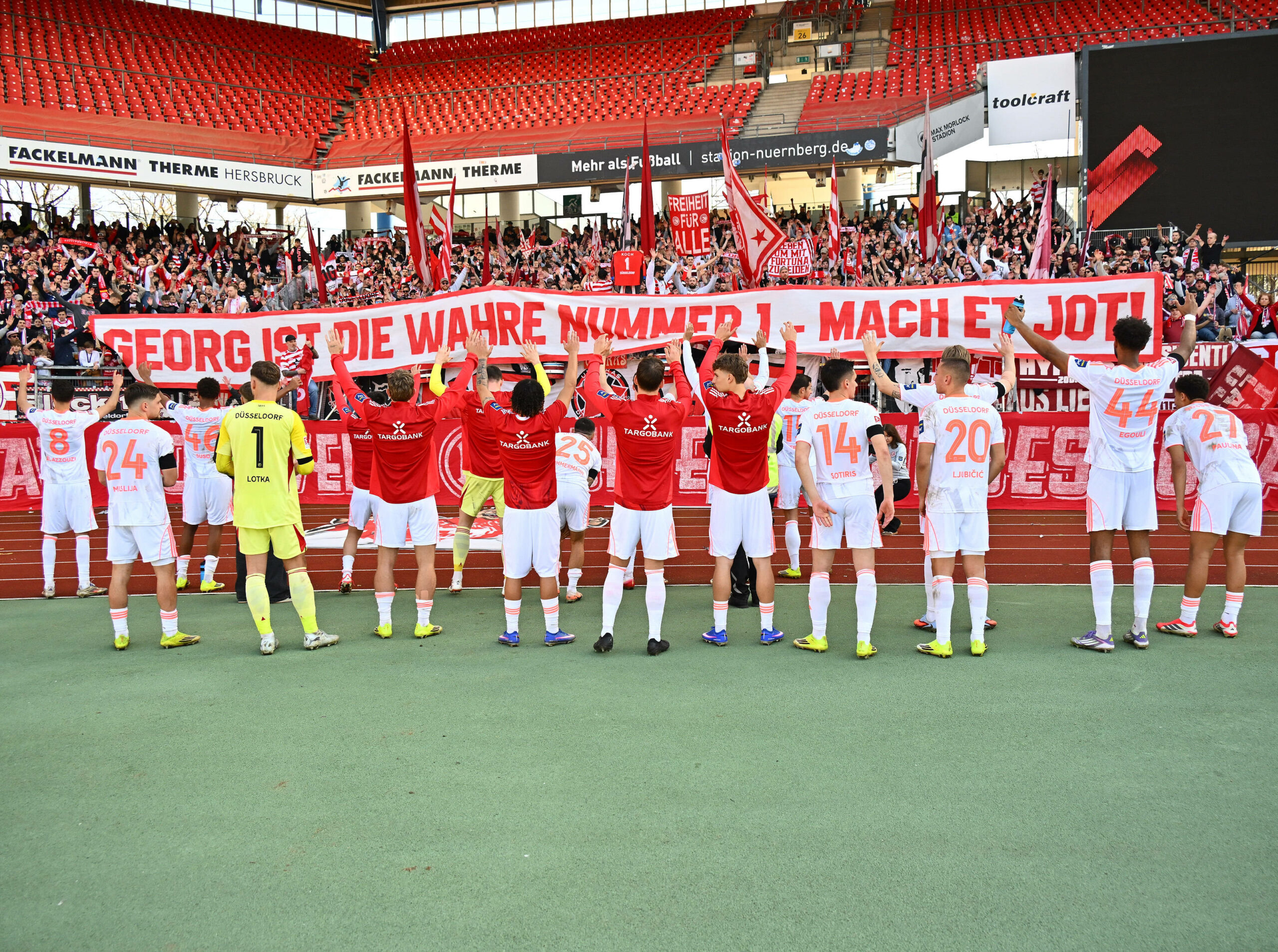 Die Mannschaft von Fortuna Düsseldorf vor den Fans mit einem Banner zu Ehren von Georg Koch