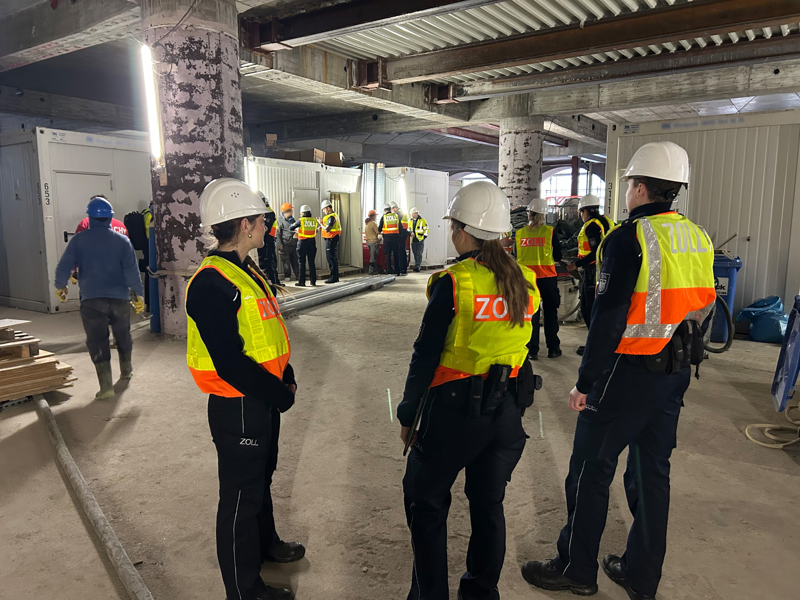 Beamte des Zolls stehen auf einer Baustelle und befragen Arbeiter.