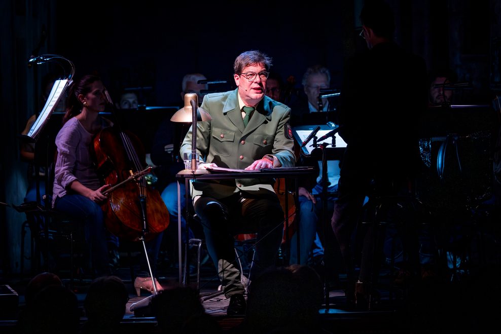 Auf der dunklen Bühne ahnt man Orchestermusiker, im Lichtschein sitzt ein Mann im Polizeikostüm an einem Tisch