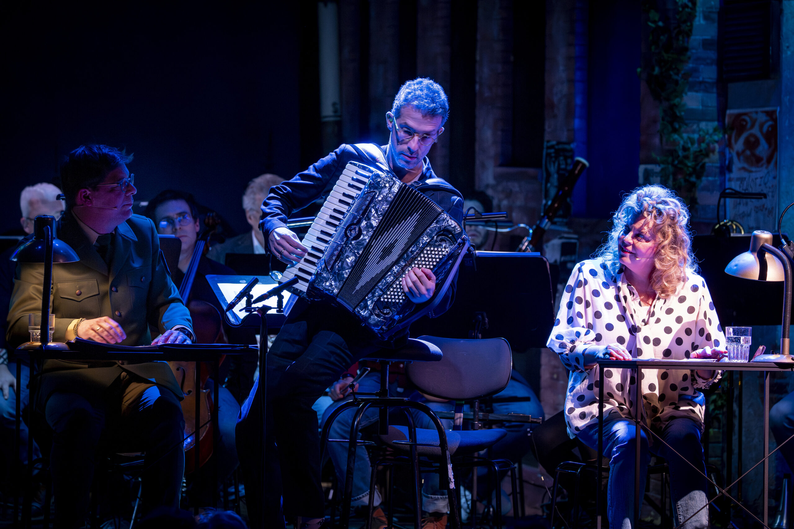 Im Hintergrund im Dunkeln das Orchester, vorne am Akkordeon Omer Meir Wellber, neben ihm sitzt an einem Tisch Carolin Spieß und sieht ihn an