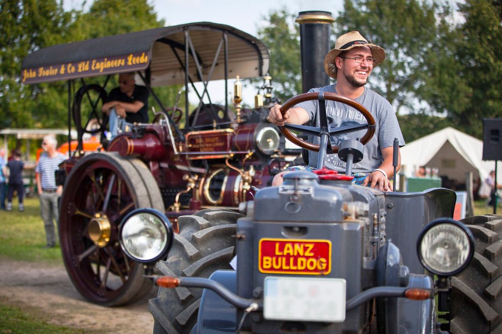 Ein Mann fährt einen historischen Lanz-Bulldog-Traktor, im Hintergrund ist eine große dampfbetriebene Maschine bei einem Oldtimer-Treffen zu sehen.