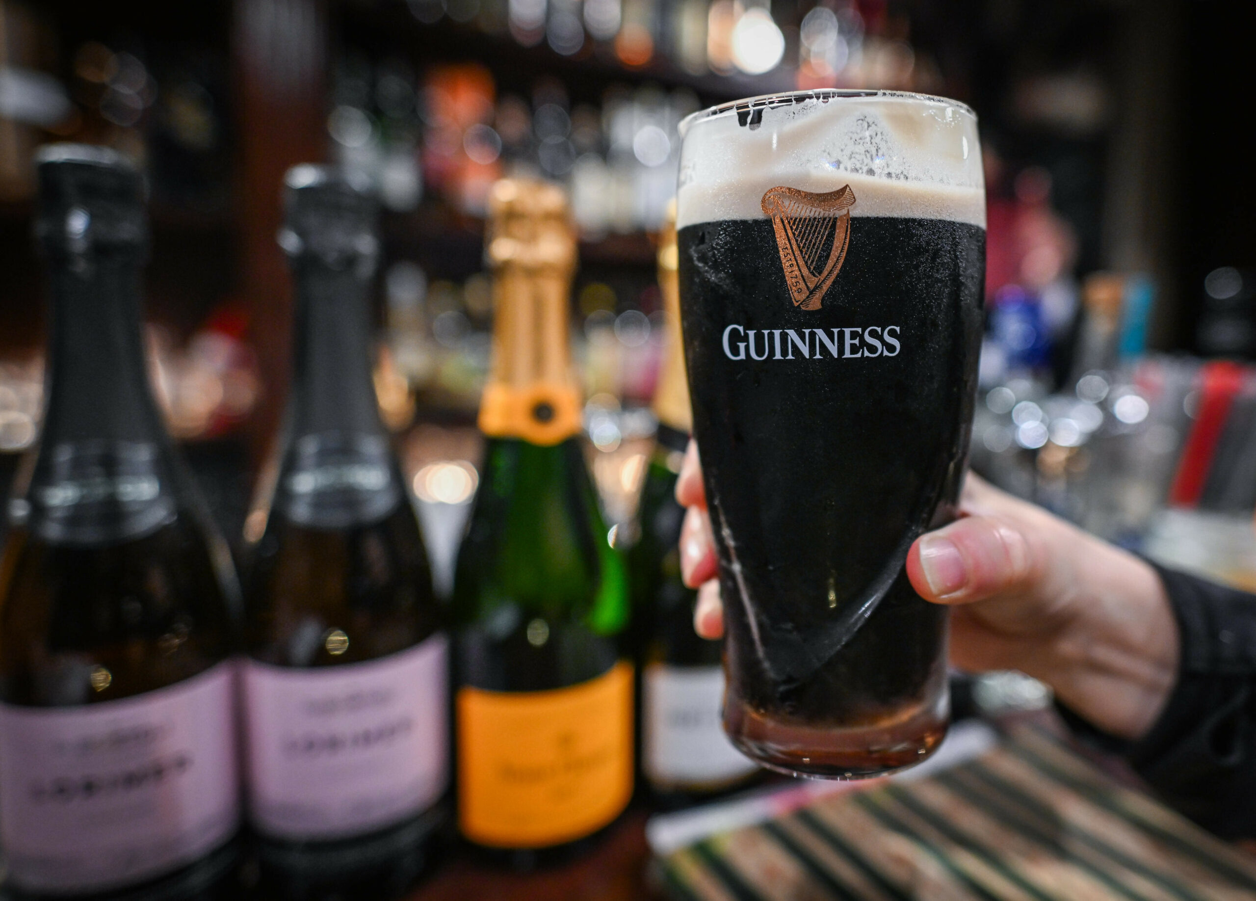 Hand hält ein Glas dunkles Bier (Guinness) vor einer Bar mit Flaschen im Hintergrund