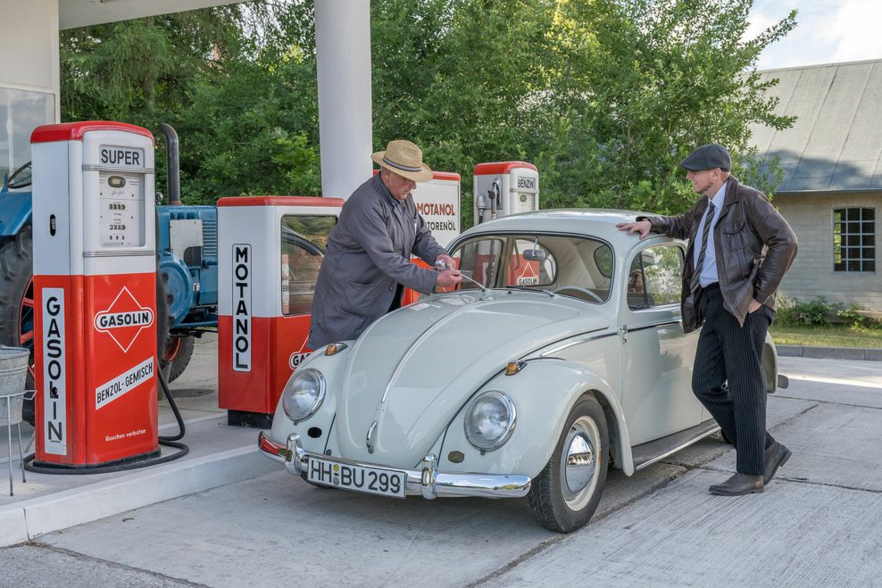 Zwei Männer in historischer Kleidung stehen an einer alten Tankstelle und betanken einen weißen VW Käfer bei einem Oldtimer-Event.