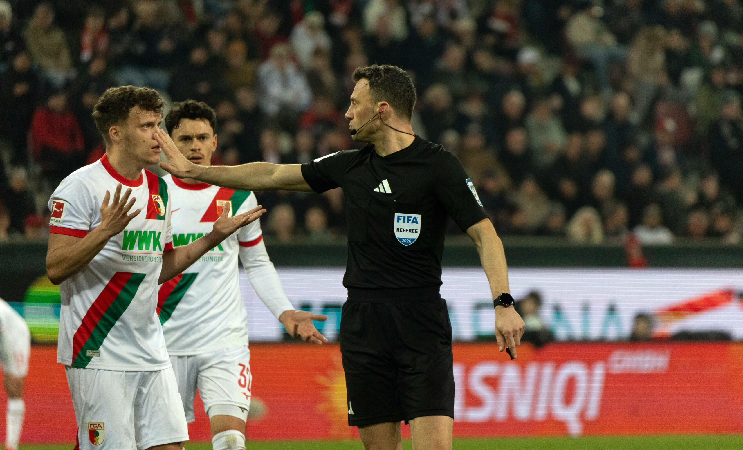 Zwayer fordert Augsburger mit ausgeschreckter Hand zum Schweigen auf.