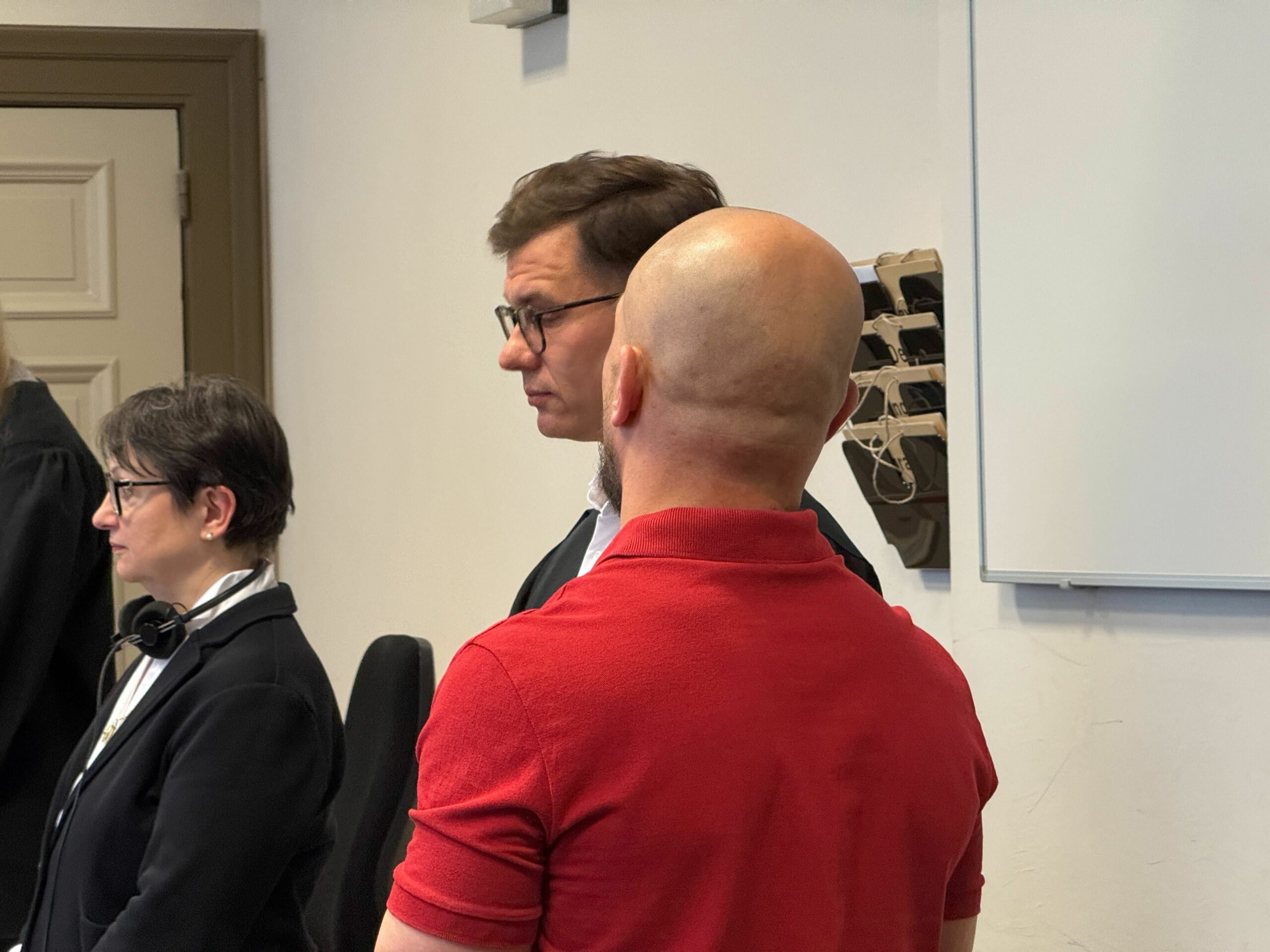 Der Angeklagte trägt ein rotes Kurzarm-Shirt und steht mit dem Rücken zur Kamera.