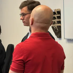Der Angeklagte trägt ein rotes Kurzarm-Shirt und steht mit dem Rücken zur Kamera.
