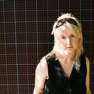 Sie steht vor einer Wand, Sonnenbrille im Haar und schaut in die Kamera.