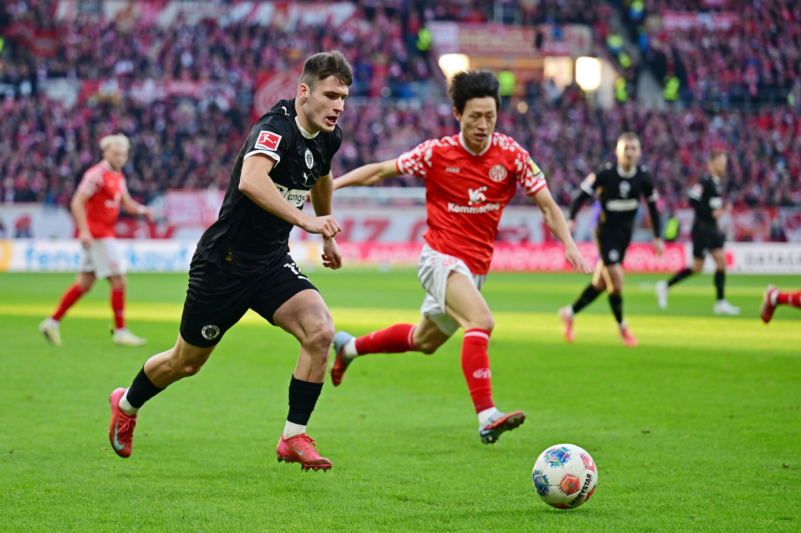 Arkadiusz Pyrka von St. Pauli im Zweikampf mit Jae-Sung Lee von Mainz vor einer TV-Kamera von DAZN