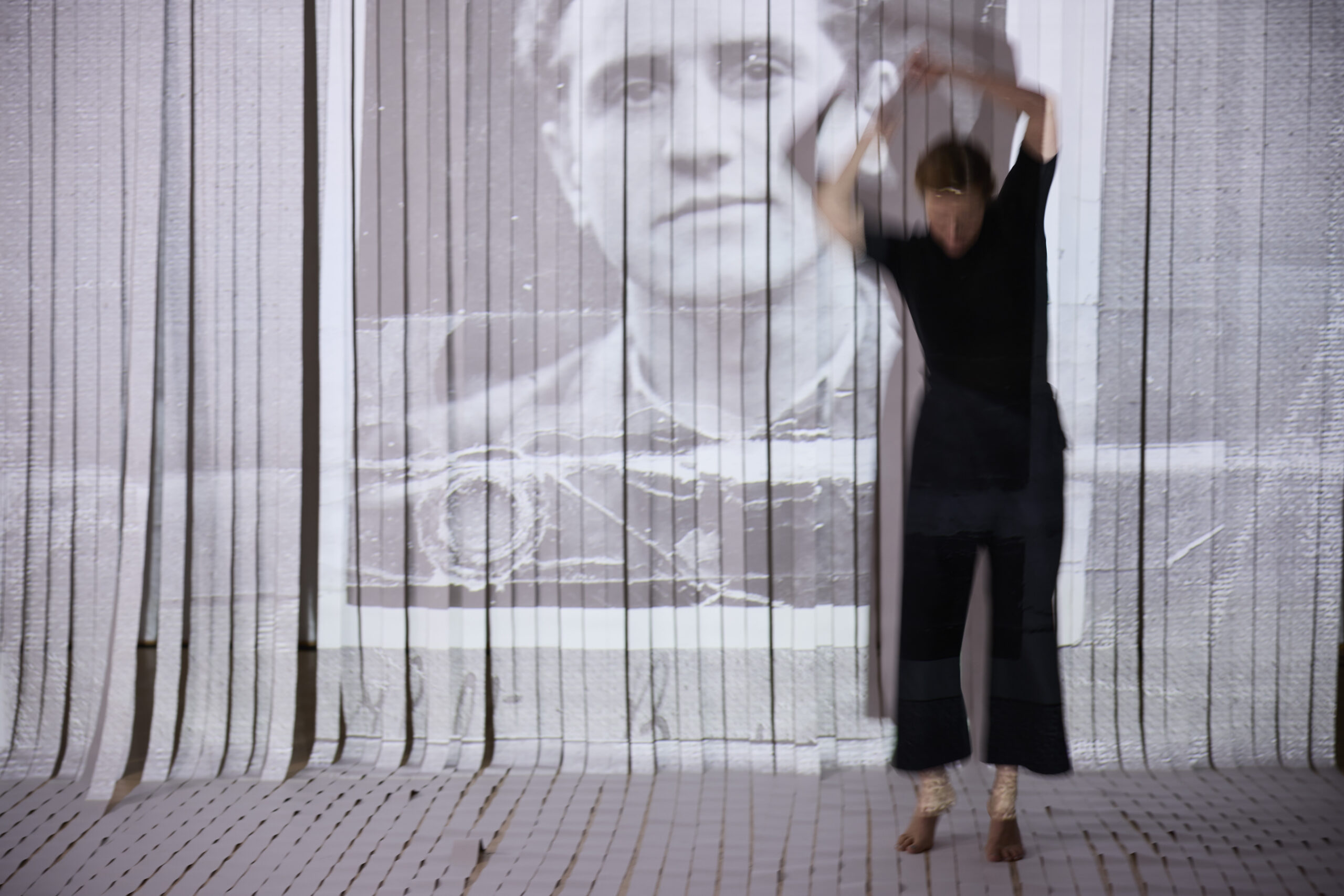 Eine Frau tanzt vor einer weißen Wand, auf welche ein Bild eines Mannes projiziert ist.