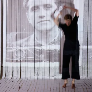 Eine Frau tanzt vor einer weißen Wand, auf welche ein Bild eines Mannes projiziert ist.