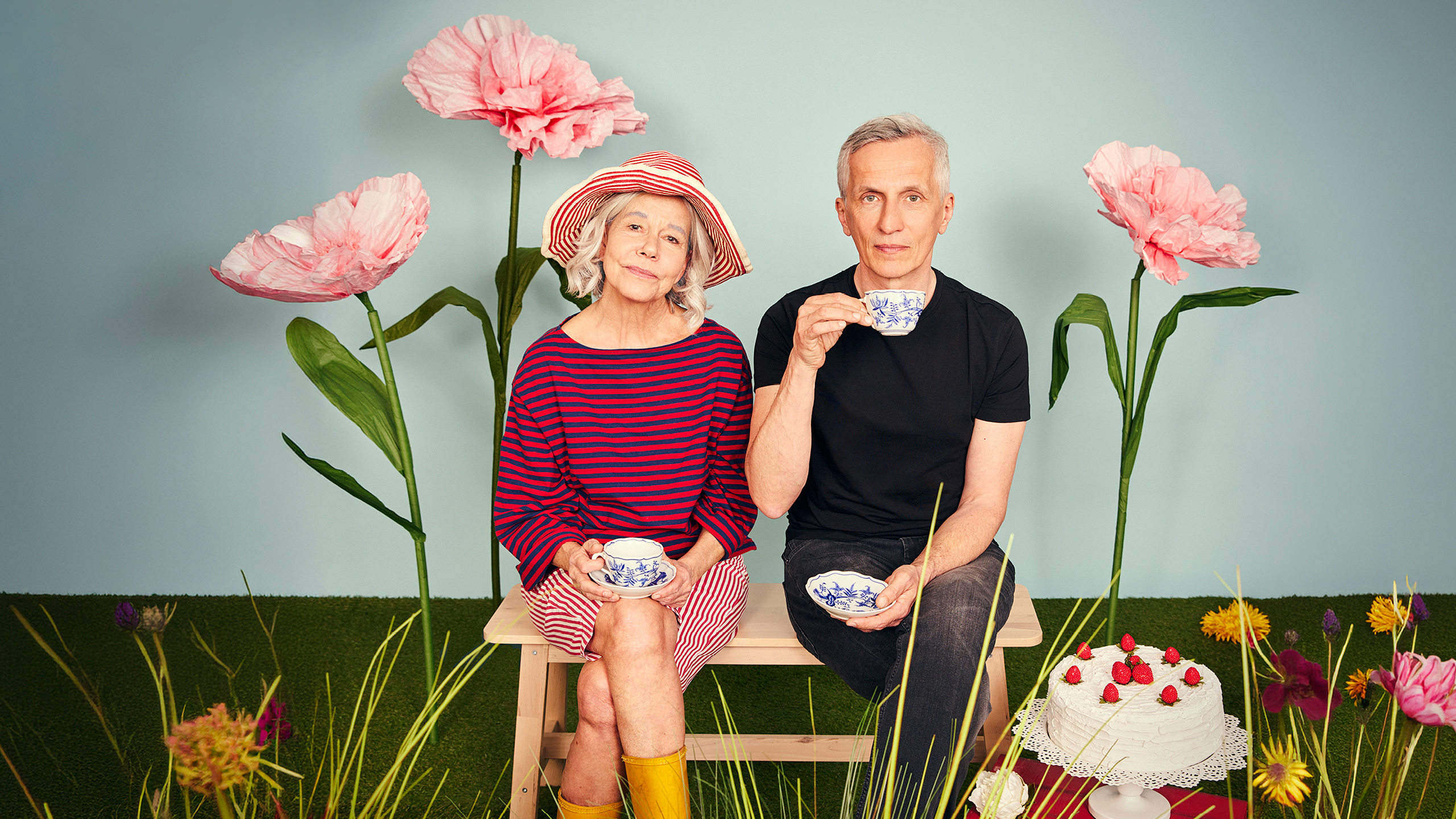 Eine Frau und ein Mann sitzen auf einer Holzbank zwischen großer Blumen.