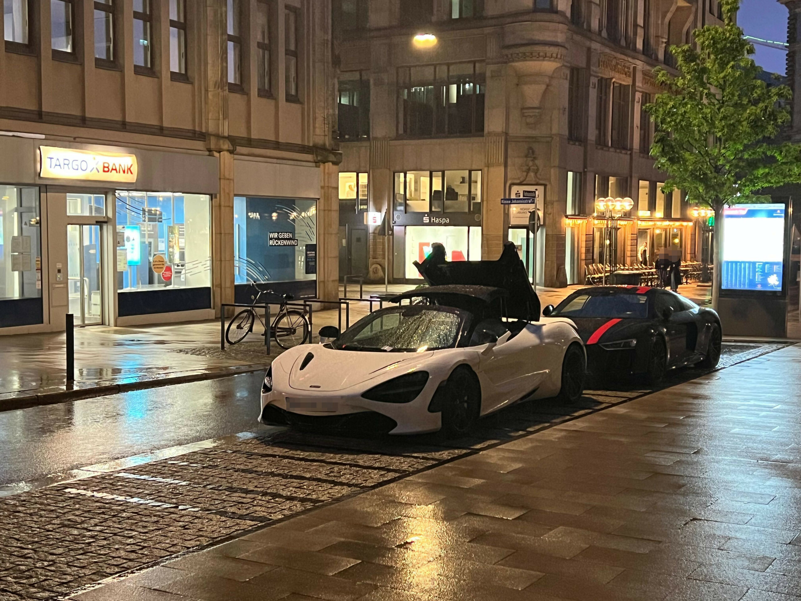 Weißer McLaren 720S Spider steht nachts bei Regen in der Hamburger Rathausstraße mit teilweise geöffnetem Verdeck am Straßenrand. Hinter dem Sportwagen parkt ein schwarzer Audi R8.